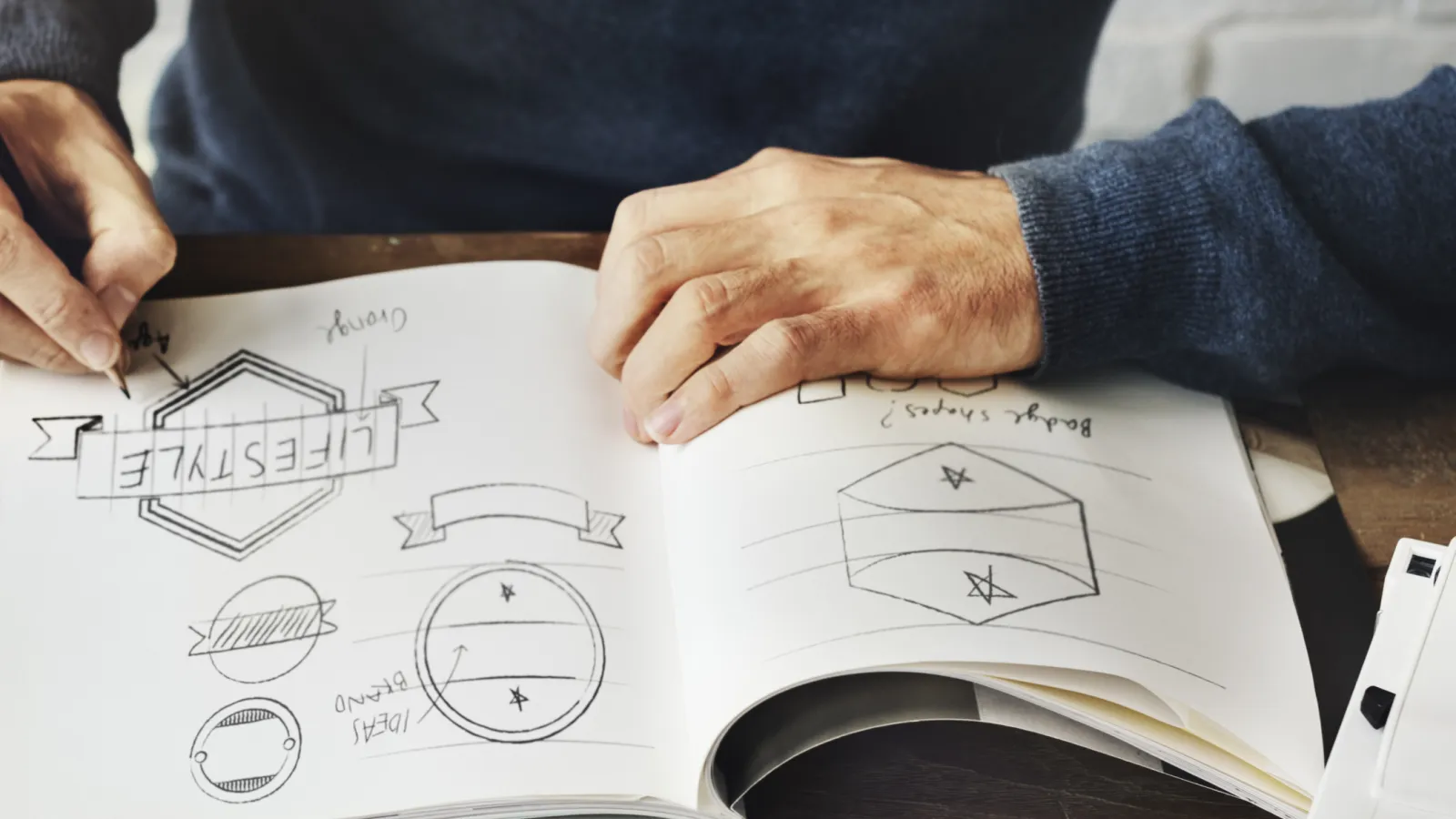Person sketching logo designs and geometric shapes in a notebook at a wooden table
