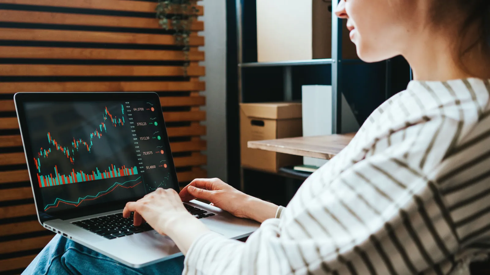 Person analyzing stock market charts and data on laptop in a modern workspace with wooden decor.