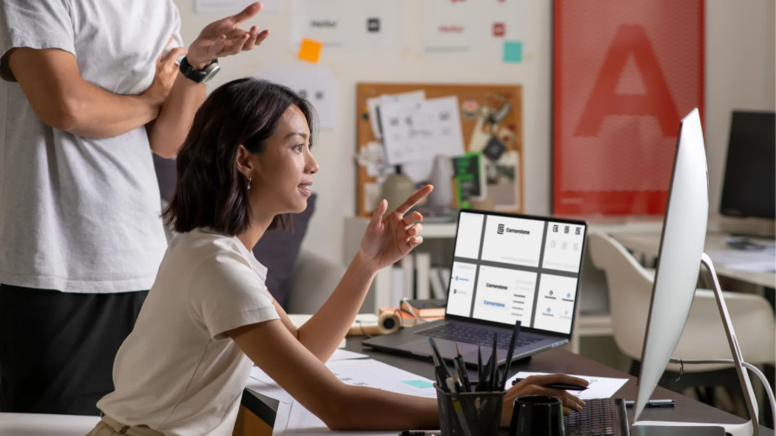 Two colleagues collaborating on a design project with a laptop and large monitor in a creative office space.