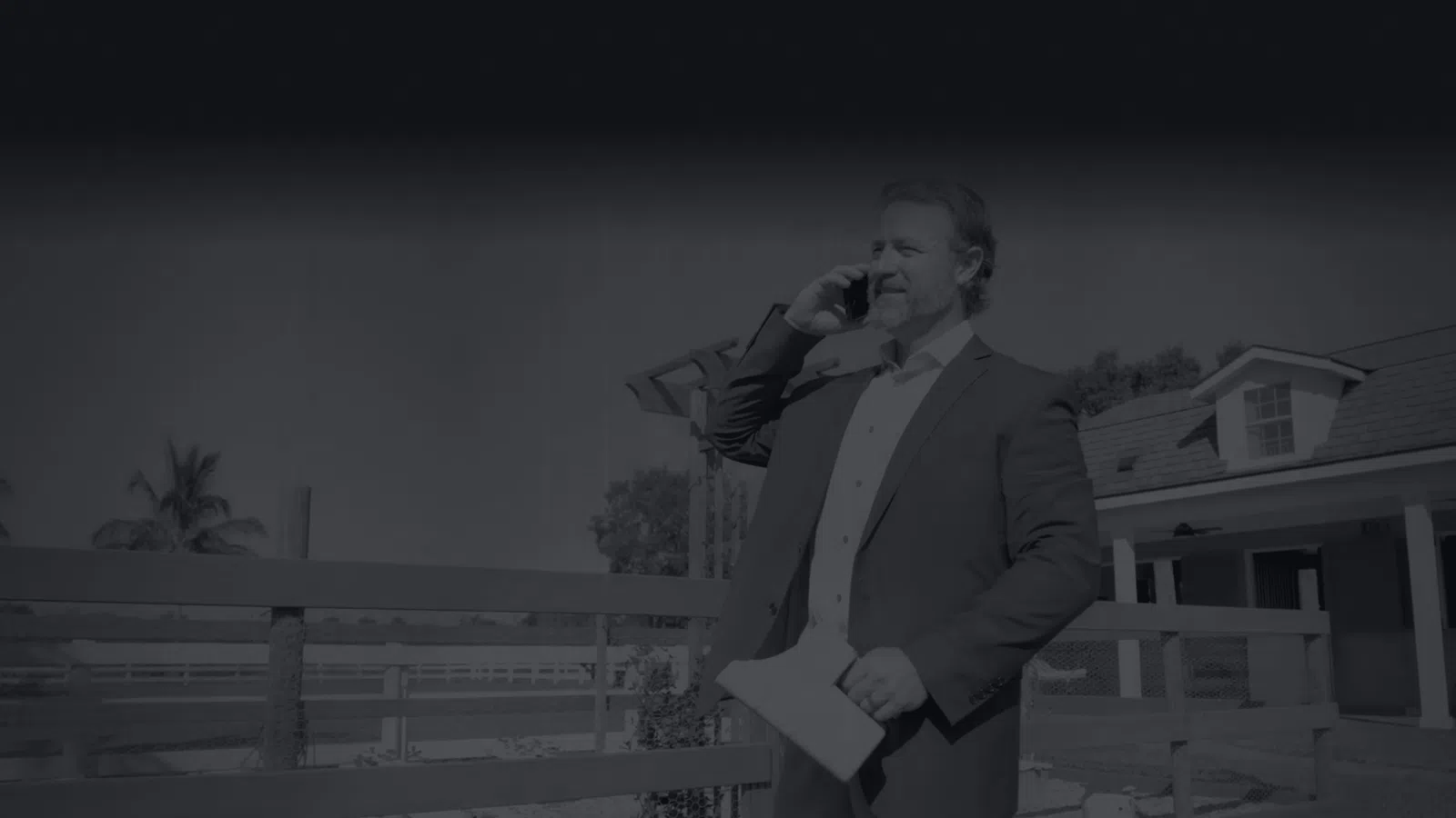 Real estate agent talking on phone holding papers standing outdoors near a house and wooden fence.