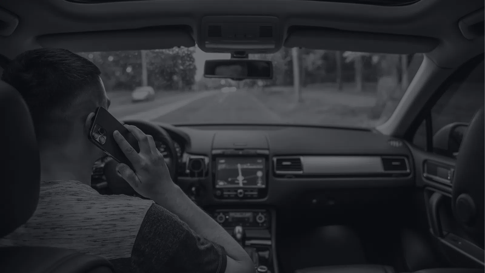 Man inside car talking on phone while driving on an empty road in daylight.