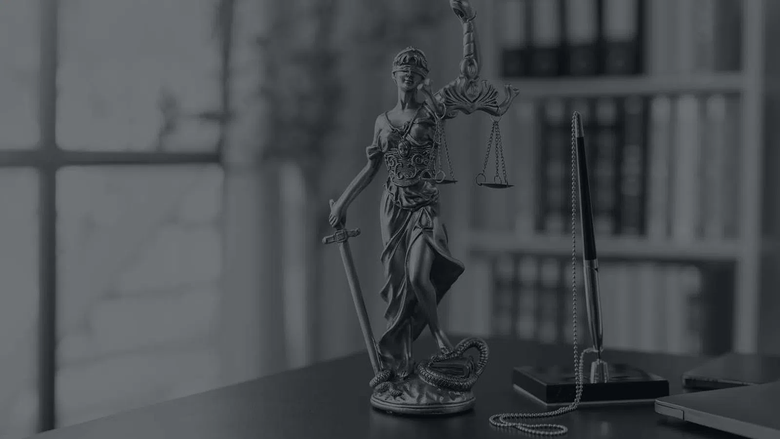 Bronze Lady Justice statue holding scales and sword on a desk with pen, notepad, and bookshelf in background.