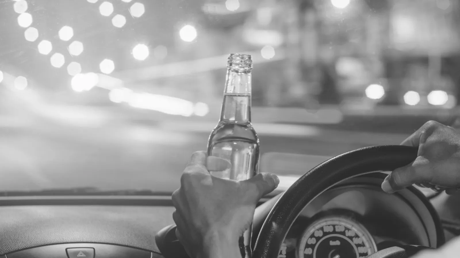Black and white image of a person holding a beer bottle while driving at night with blurred city lights ahead.
