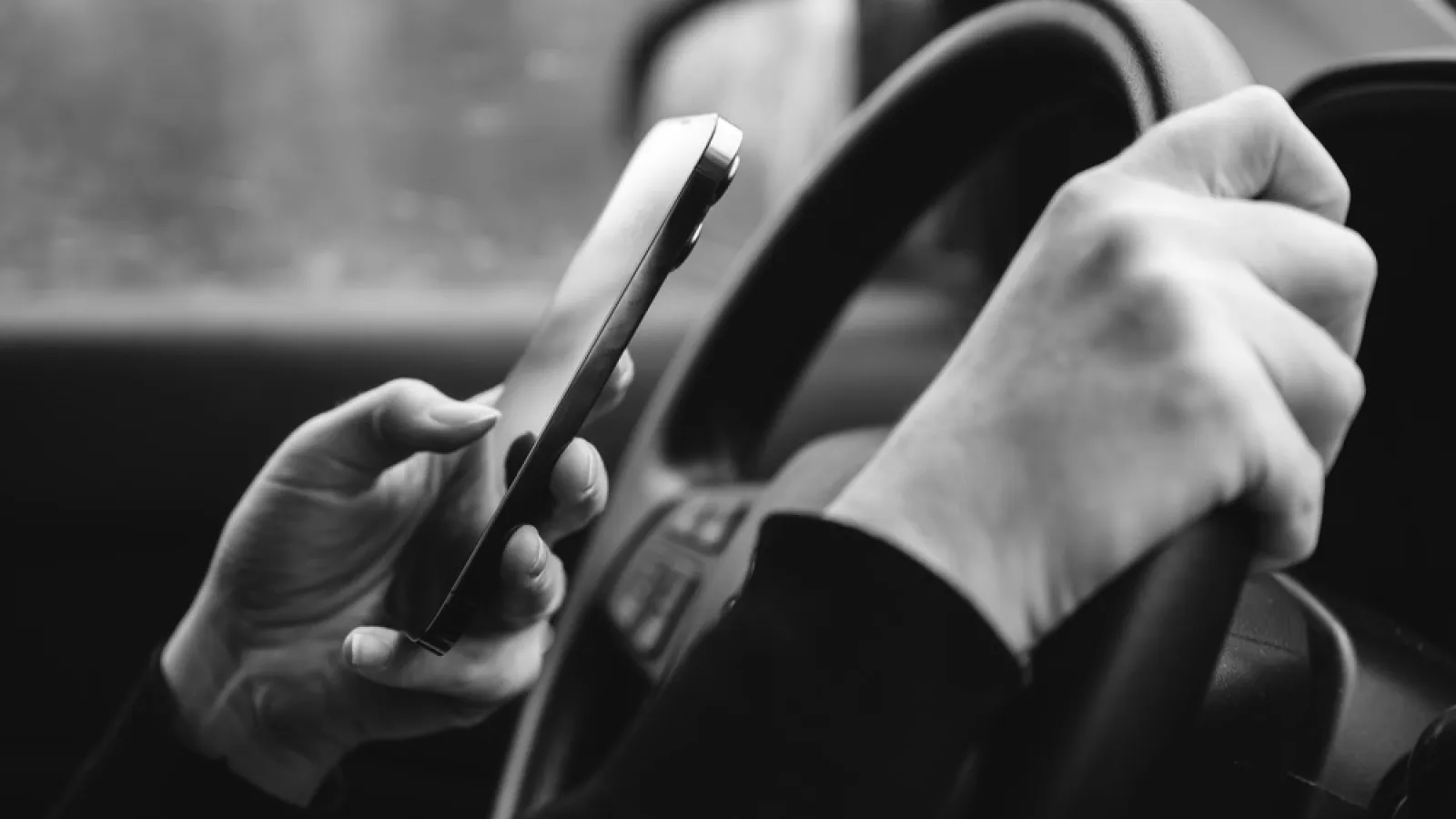 Black and white photo of a person using a smartphone while driving, one hand on the steering wheel.