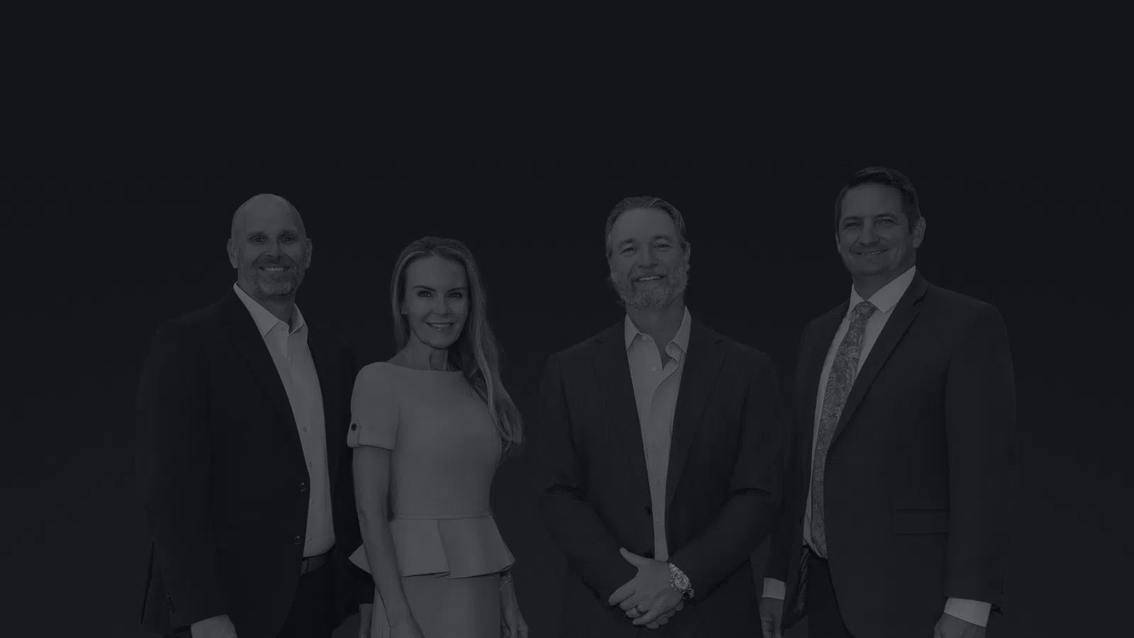 Four professionally dressed businesspeople standing side by side against a plain background, smiling.