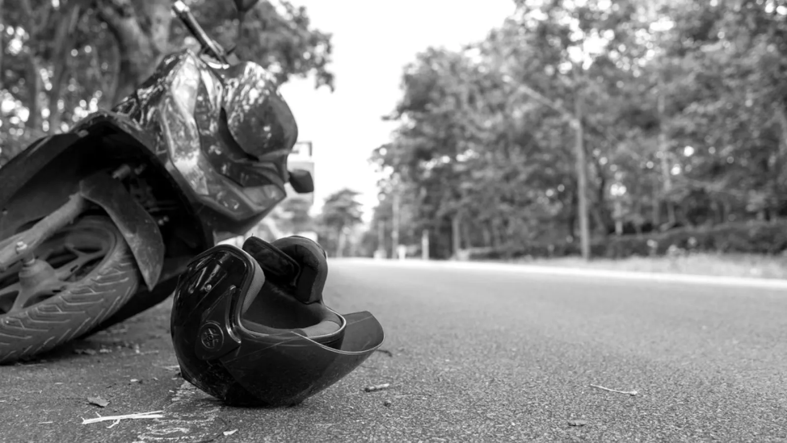 Black and white image of a fallen motorcycle and helmet on a deserted road with trees in the background