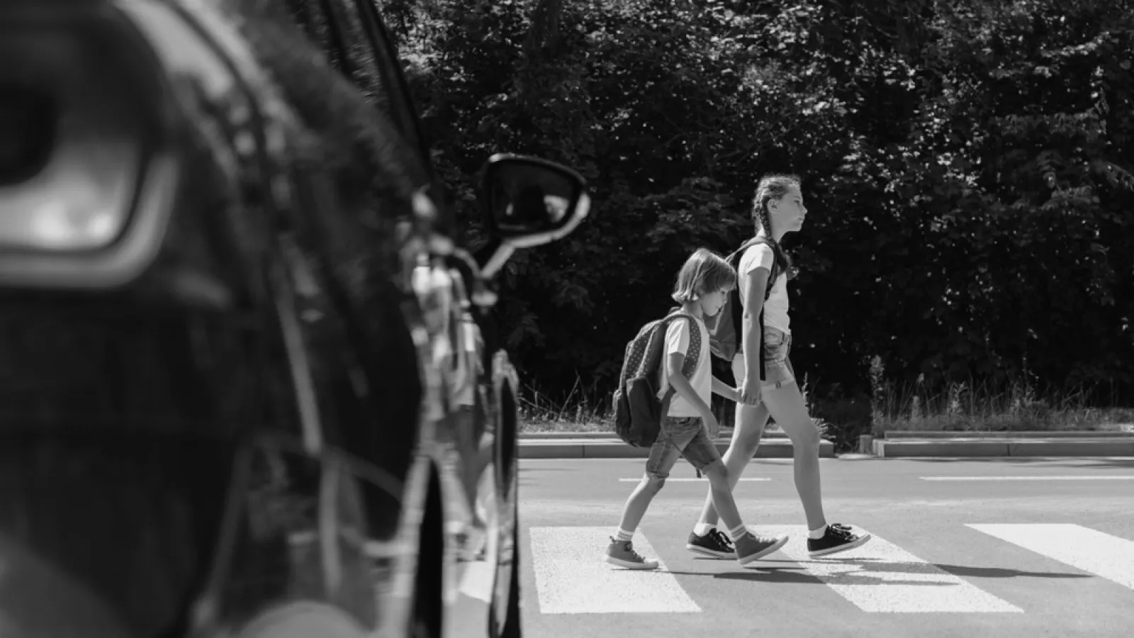 Two children with backpacks cross a street at a pedestrian crosswalk, with a car waiting nearby.