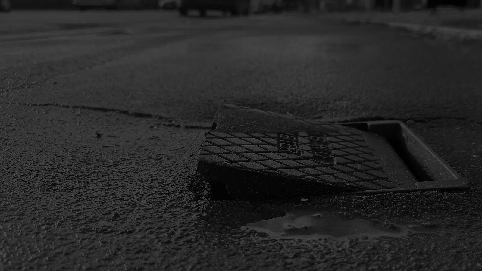 Broken storm drain cover displaced on rough asphalt street with blurred vehicles and buildings in background.