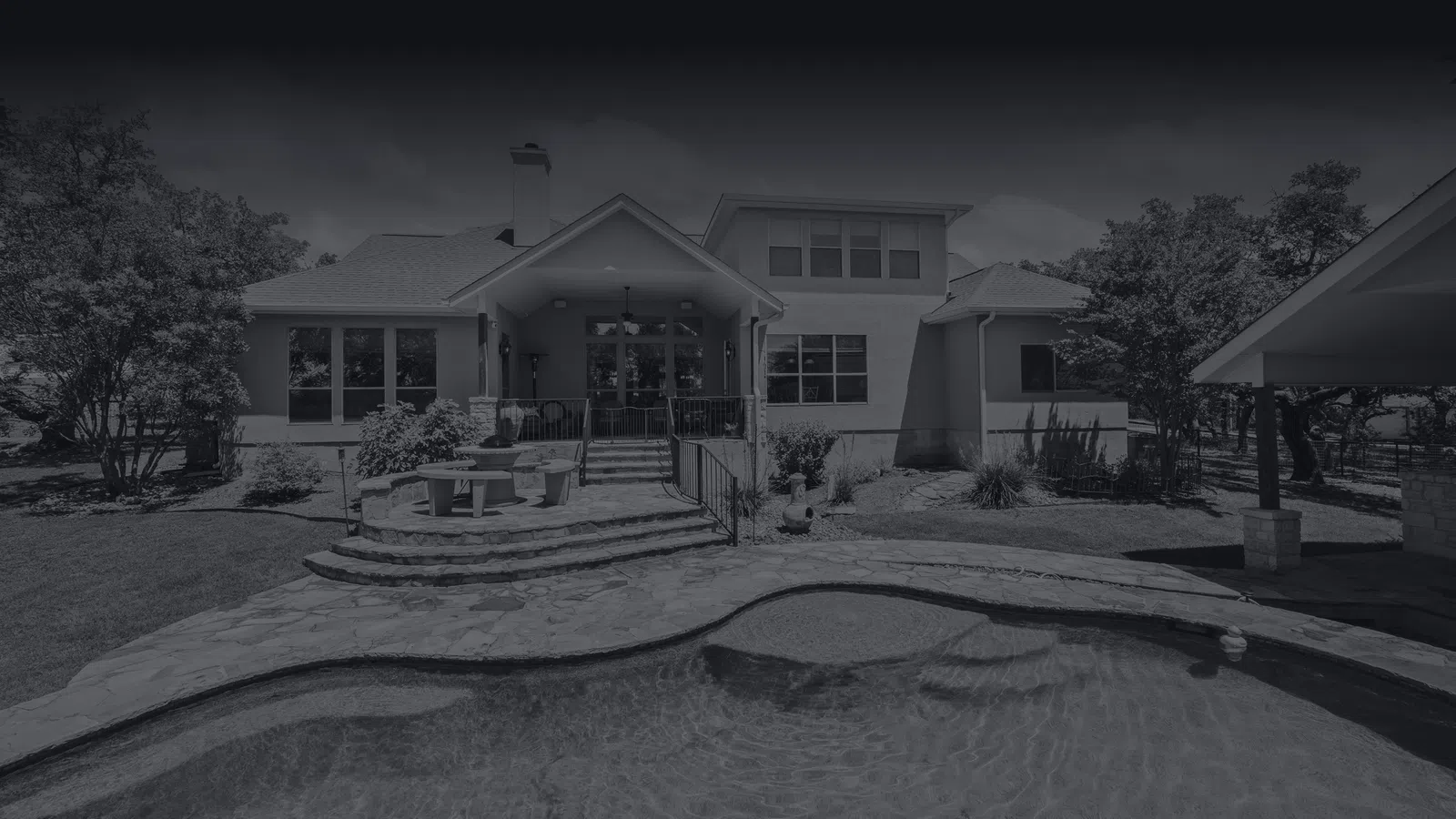 Modern house exterior with a swimming pool, stone patio, outdoor seating, and surrounding trees in daylight.