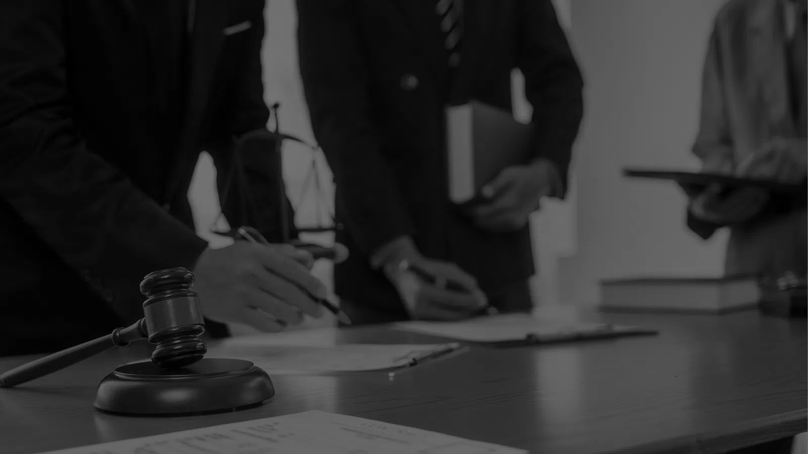 Gavel and legal documents on table with lawyers in suits discussing cases in background in black and white
