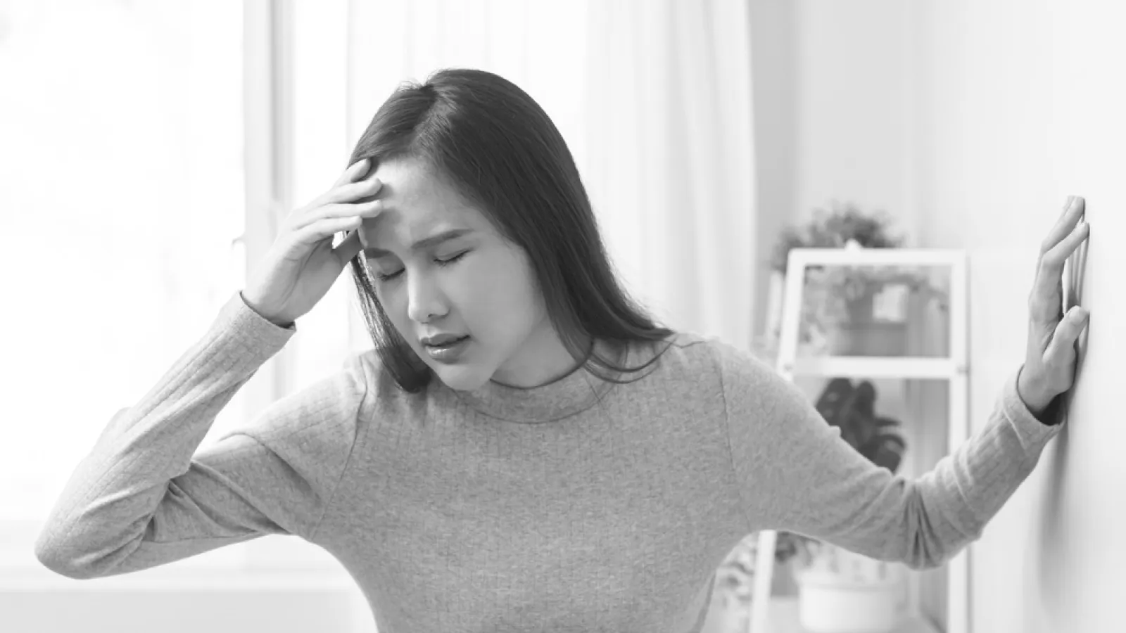 Young woman in gray sweater holding her head and leaning on wall showing signs of dizziness or headache indoors.