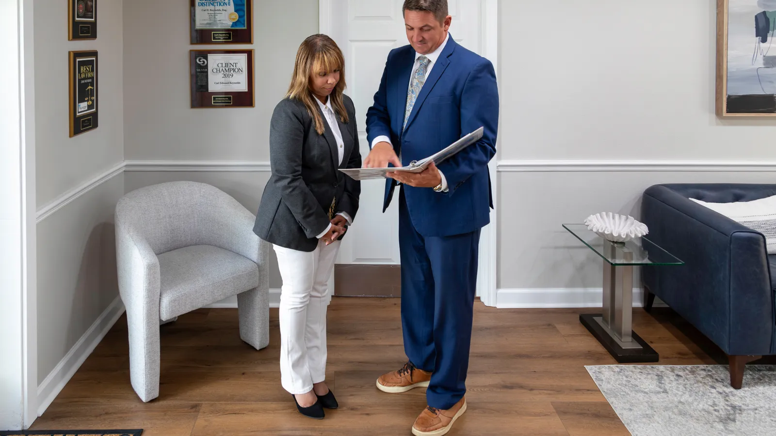 Two business professionals in suits review a document together in a modern office with awards on the wall.