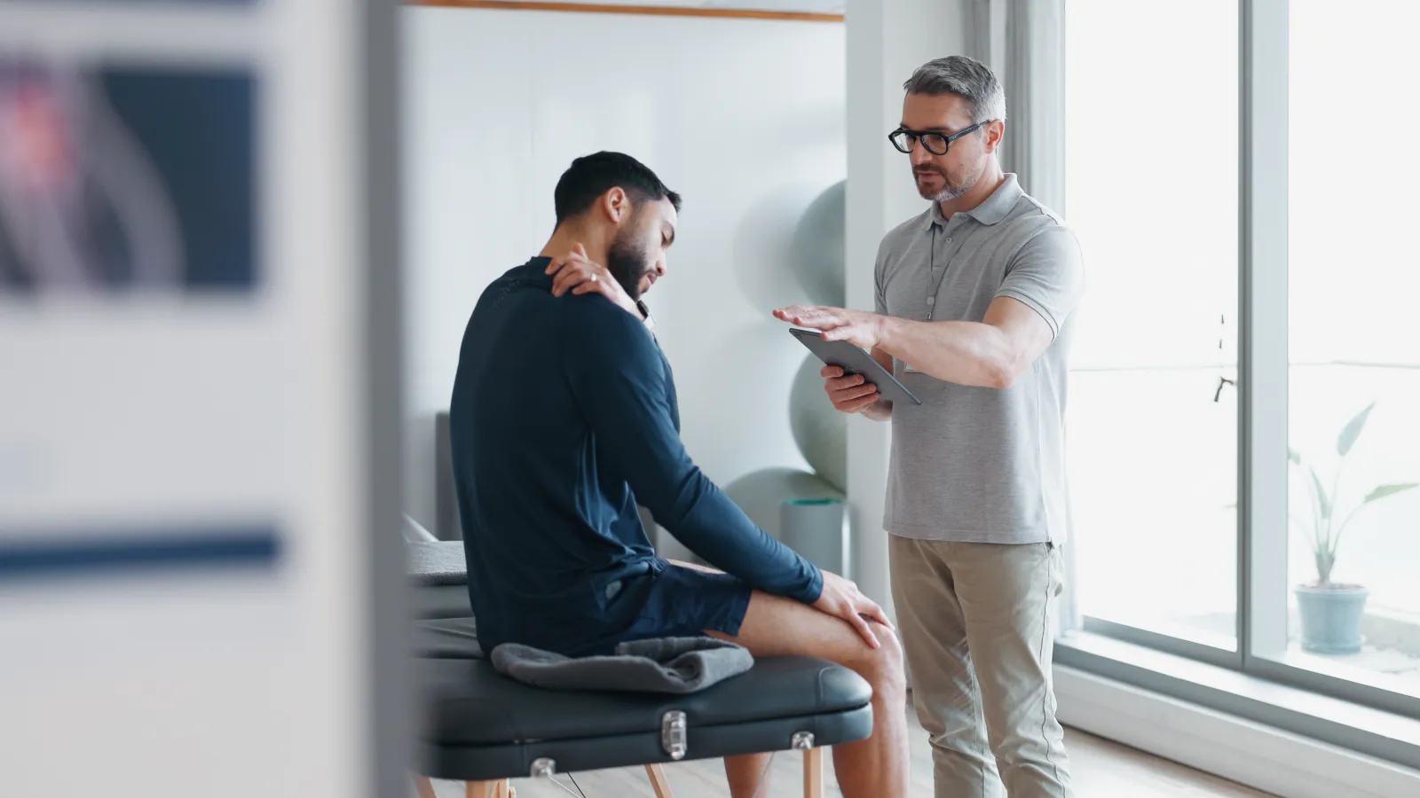 Physical therapist consulting with male patient seated on examination table in bright clinic room