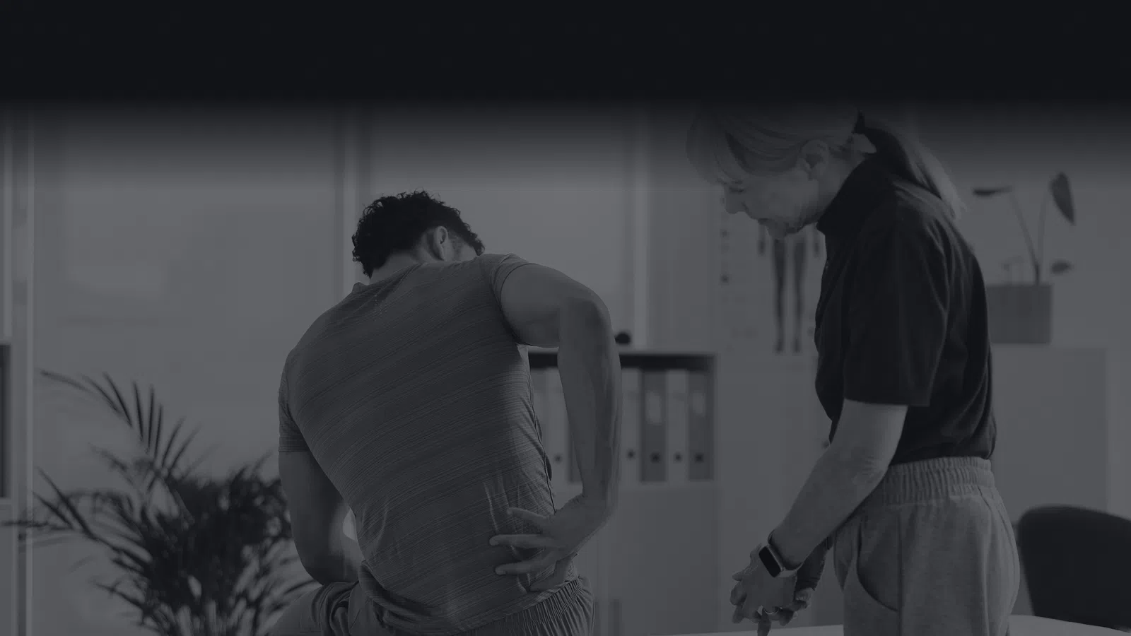 Man holding lower back in pain during physical therapy session with female therapist in clinic room.