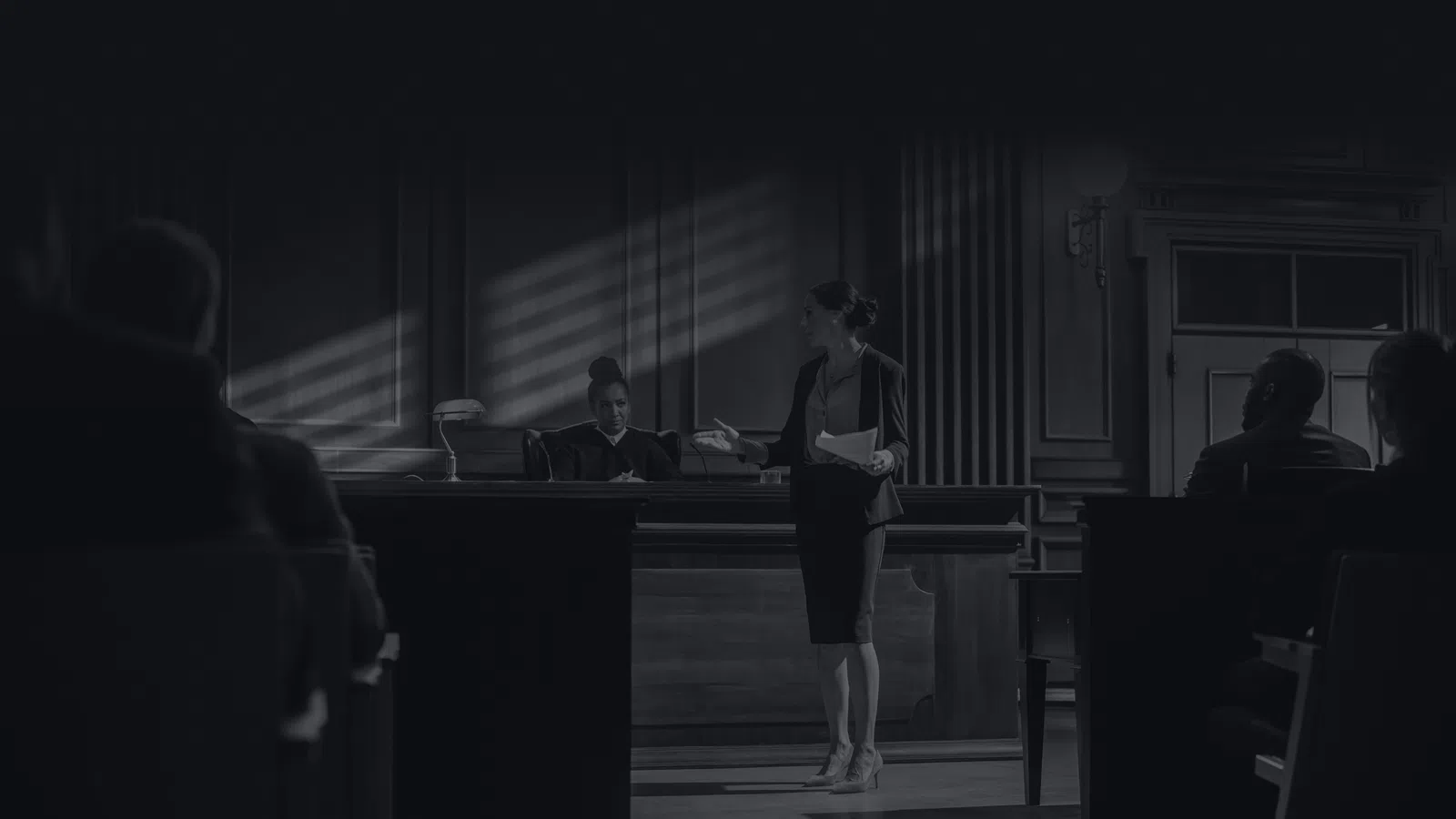 Lawyer presenting case in courtroom with judge and seated audience in black and white setting.