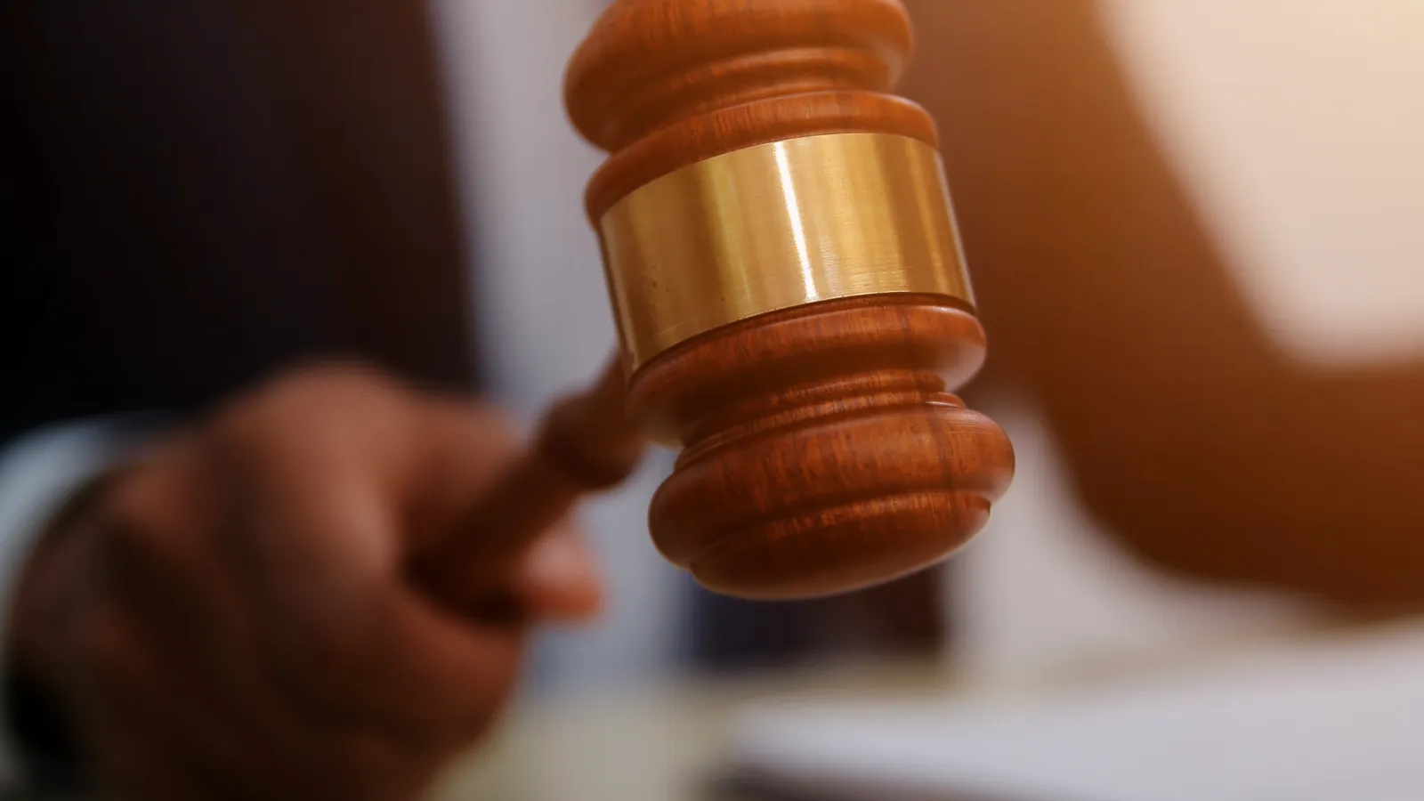 Close-up of a judge in suit striking a wooden gavel on a sound block in courtroom setting.