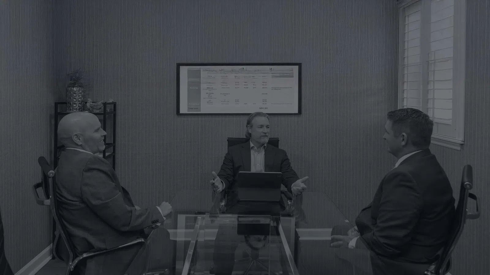 Three businessmen in suits engaged in a discussion around a glass conference table in a modern office.