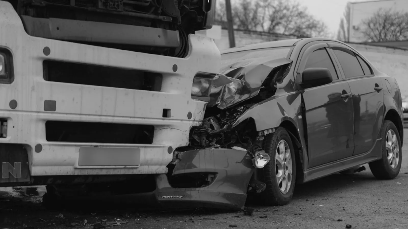 Black and white image of a car severely damaged in a collision with a large truck's front end.
