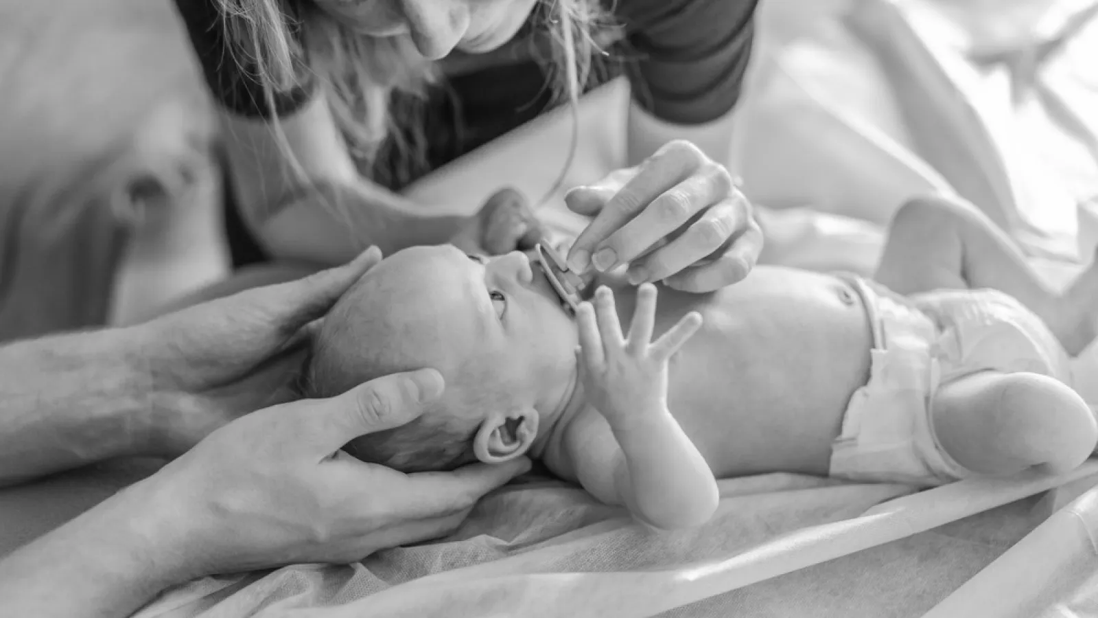 Black and white photo of a mother feeding a newborn baby with a spoon while father supports the baby’s head.