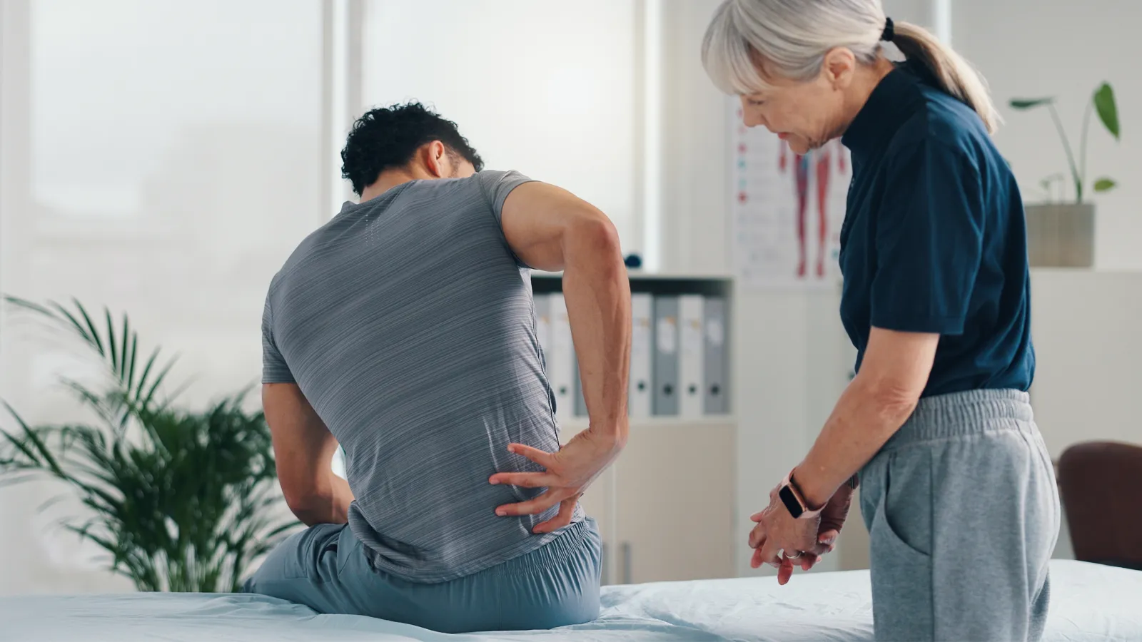 Male patient holding lower back in pain consulting elderly female doctor in a medical office clinic.