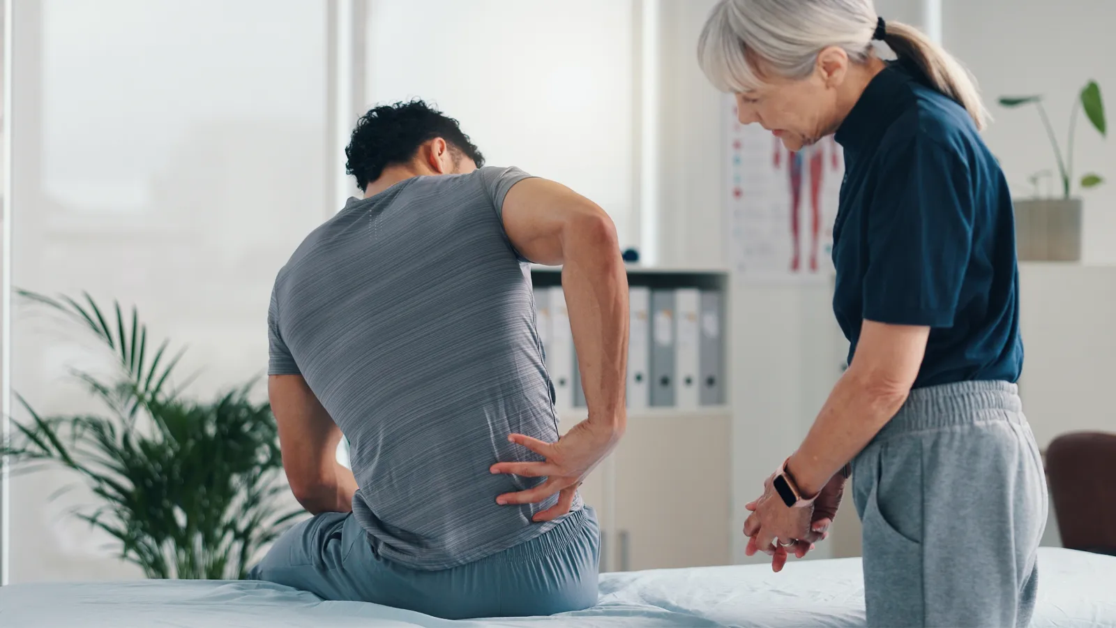 Male patient holding lower back in pain consulting elderly female doctor in a medical office clinic.