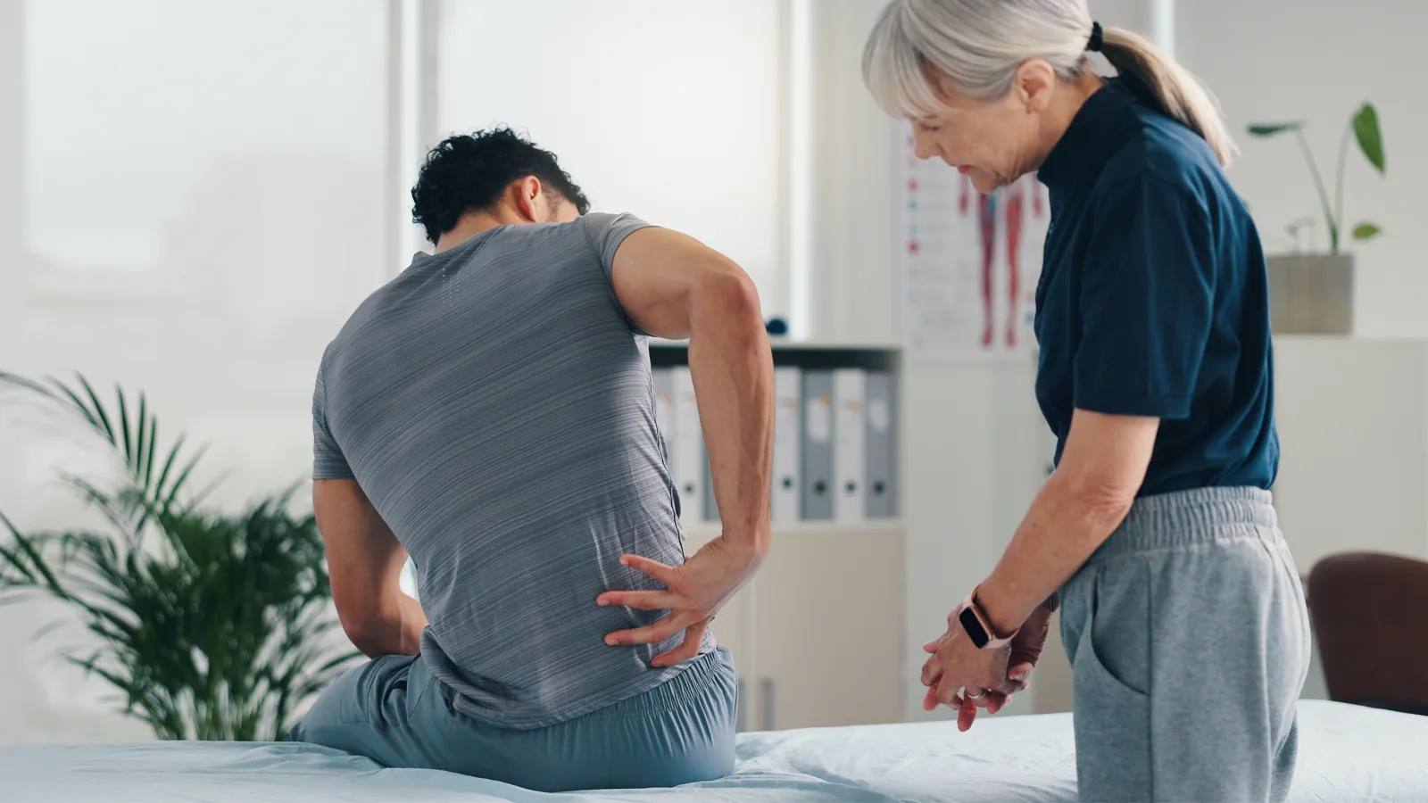 Male patient holding lower back in pain consulting elderly female doctor in a medical office clinic.