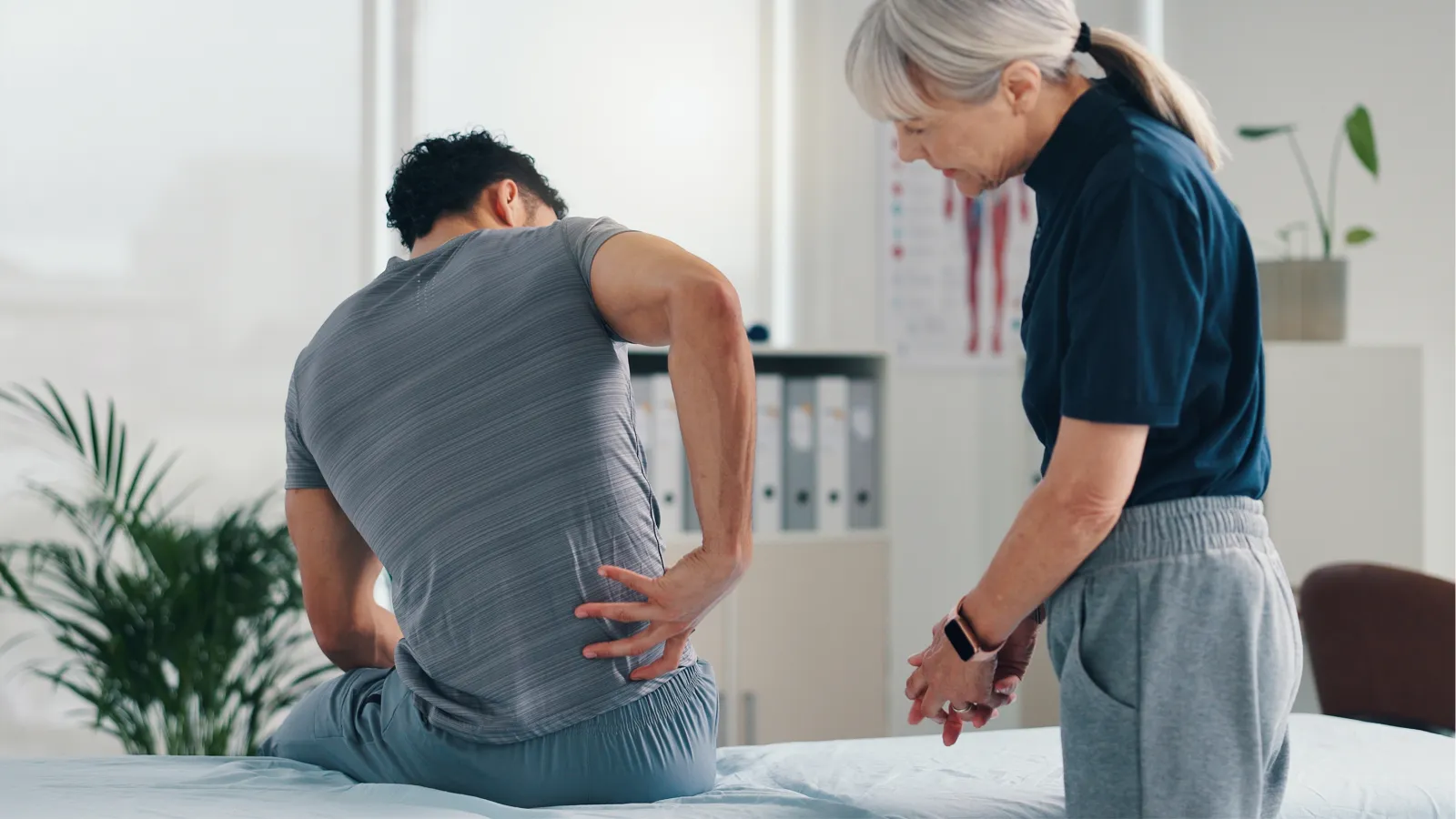 Male patient holding lower back in pain consulting elderly female doctor in a medical office clinic.