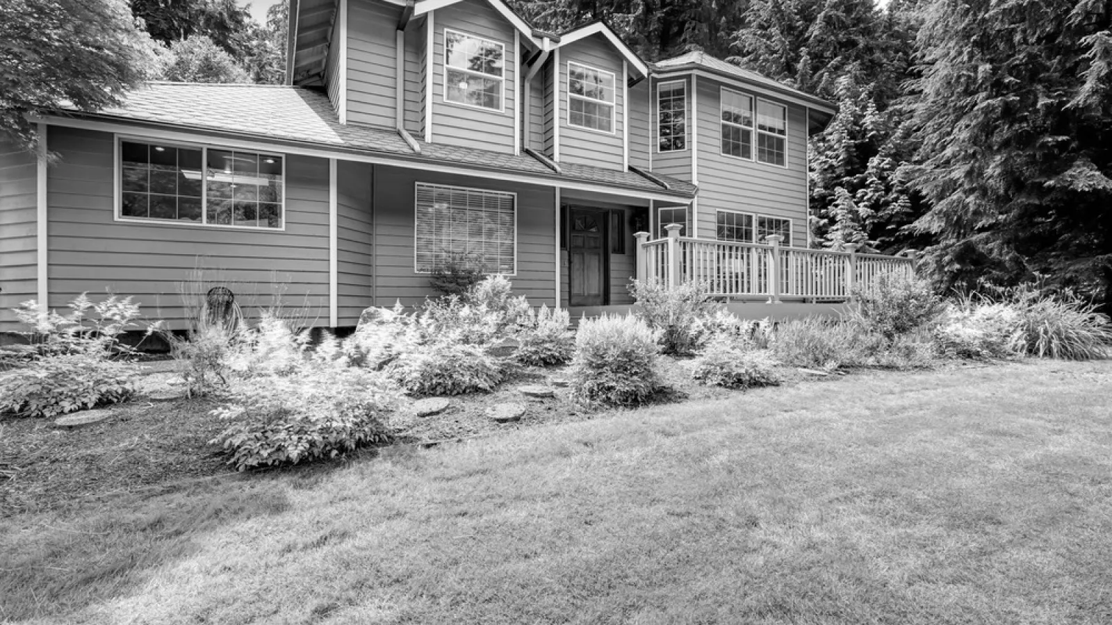 Grey two-story house with large windows, porch, and landscaped front yard surrounded by tall trees.