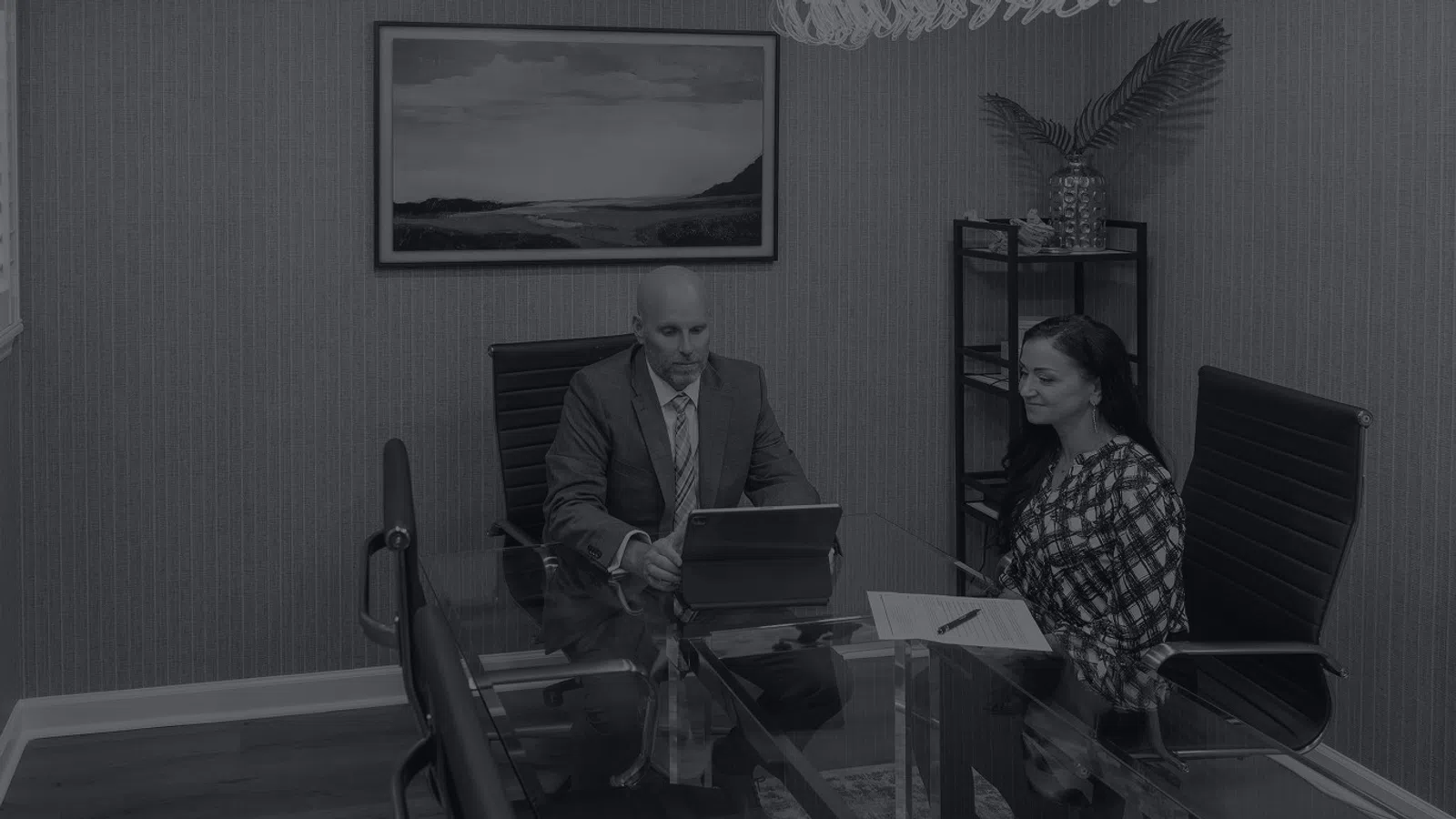 Man and woman discussing work at glass table in modern office with framed landscape and plant on shelf
