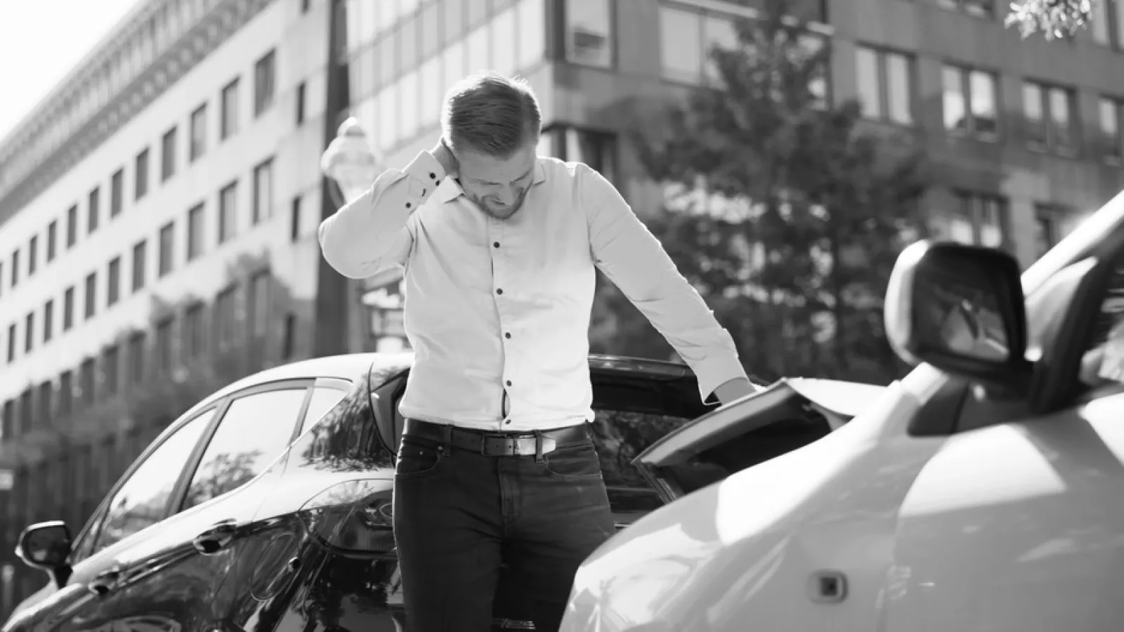 Man in white shirt looking at car damage after an accident on a city street with office buildings in background