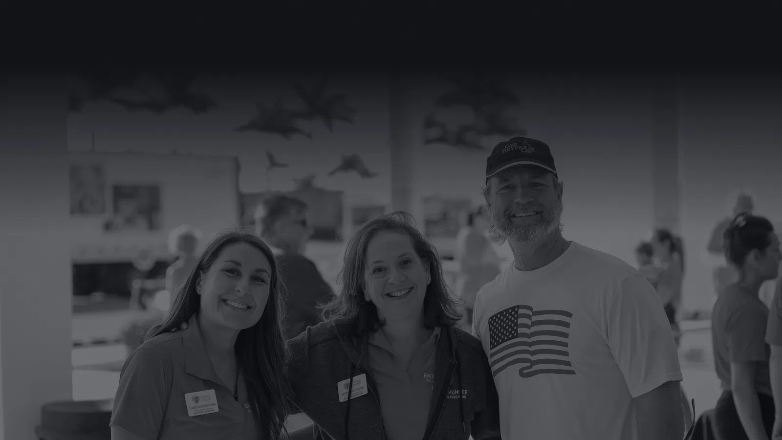 Three smiling adults posing indoors at a social or work event with blurred people in the background.
