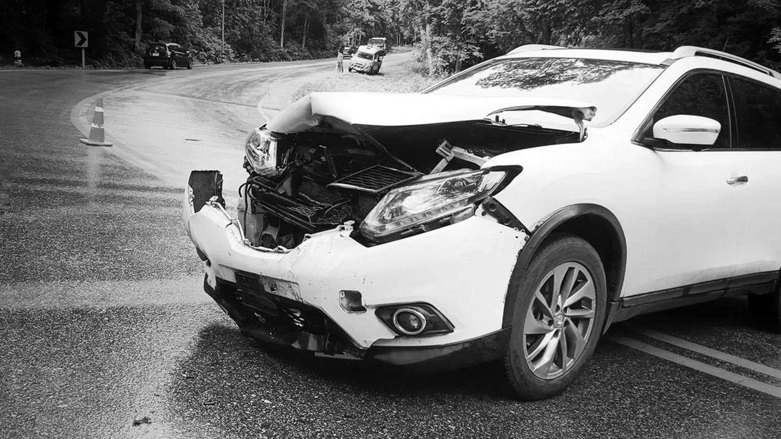 White SUV with severe front-end damage parked on a curved forest road after an accident.