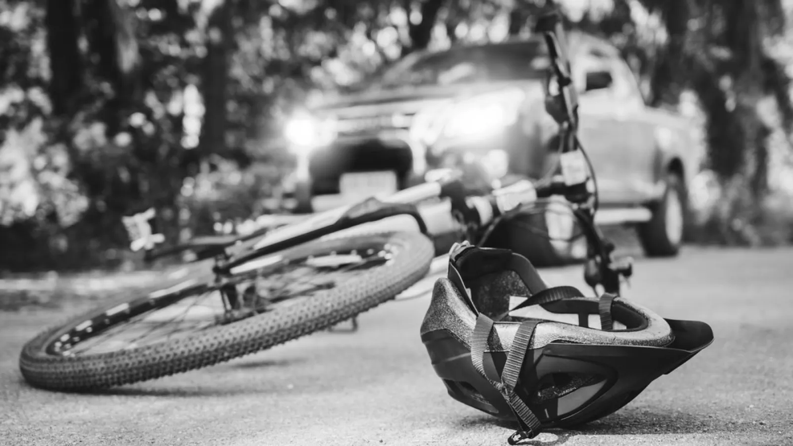 Bicycle and helmet lying on the road with a pickup truck in the background under trees.