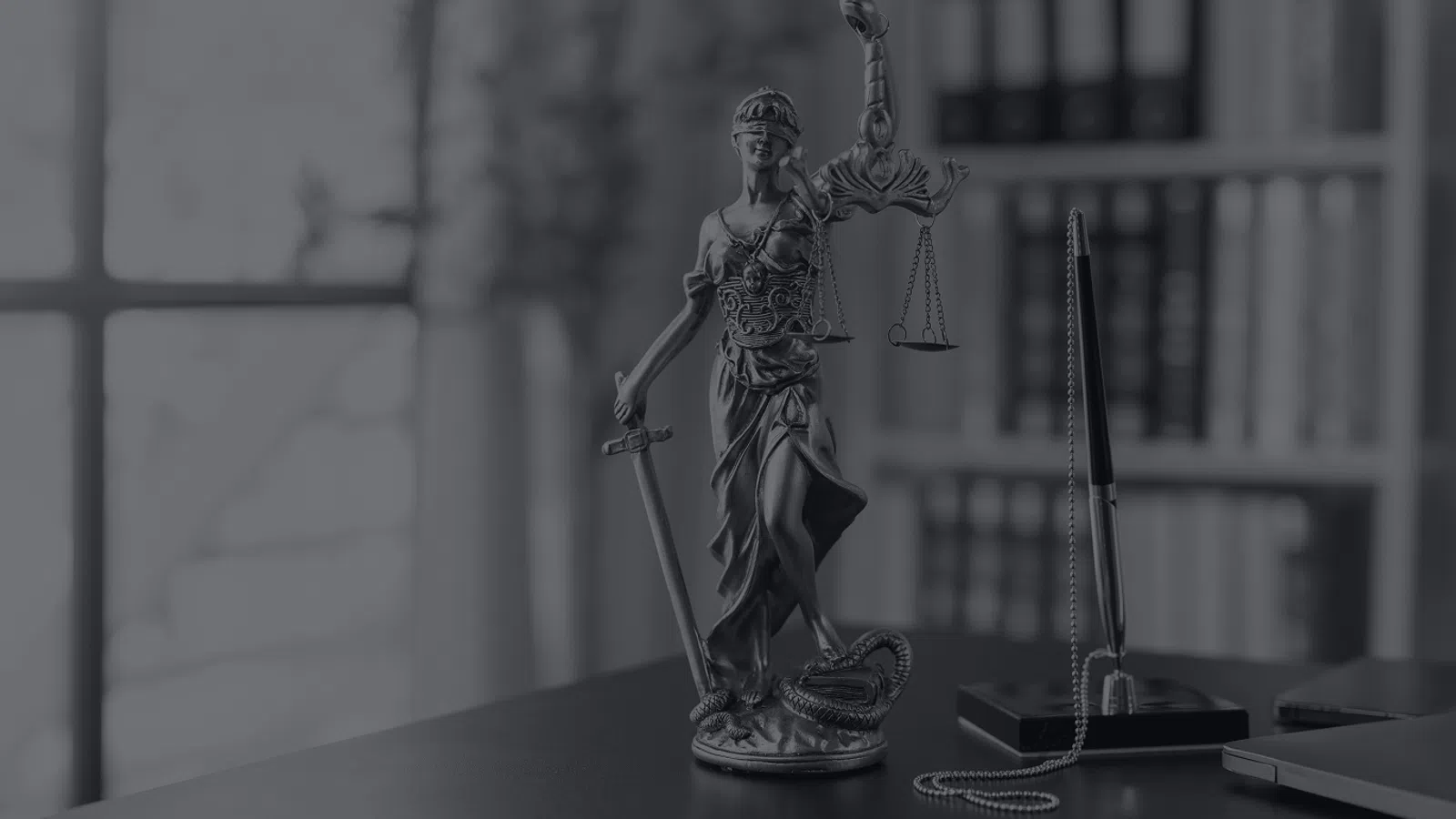 Blindfolded Lady Justice statue holding sword and scales on desk with books and pen in background