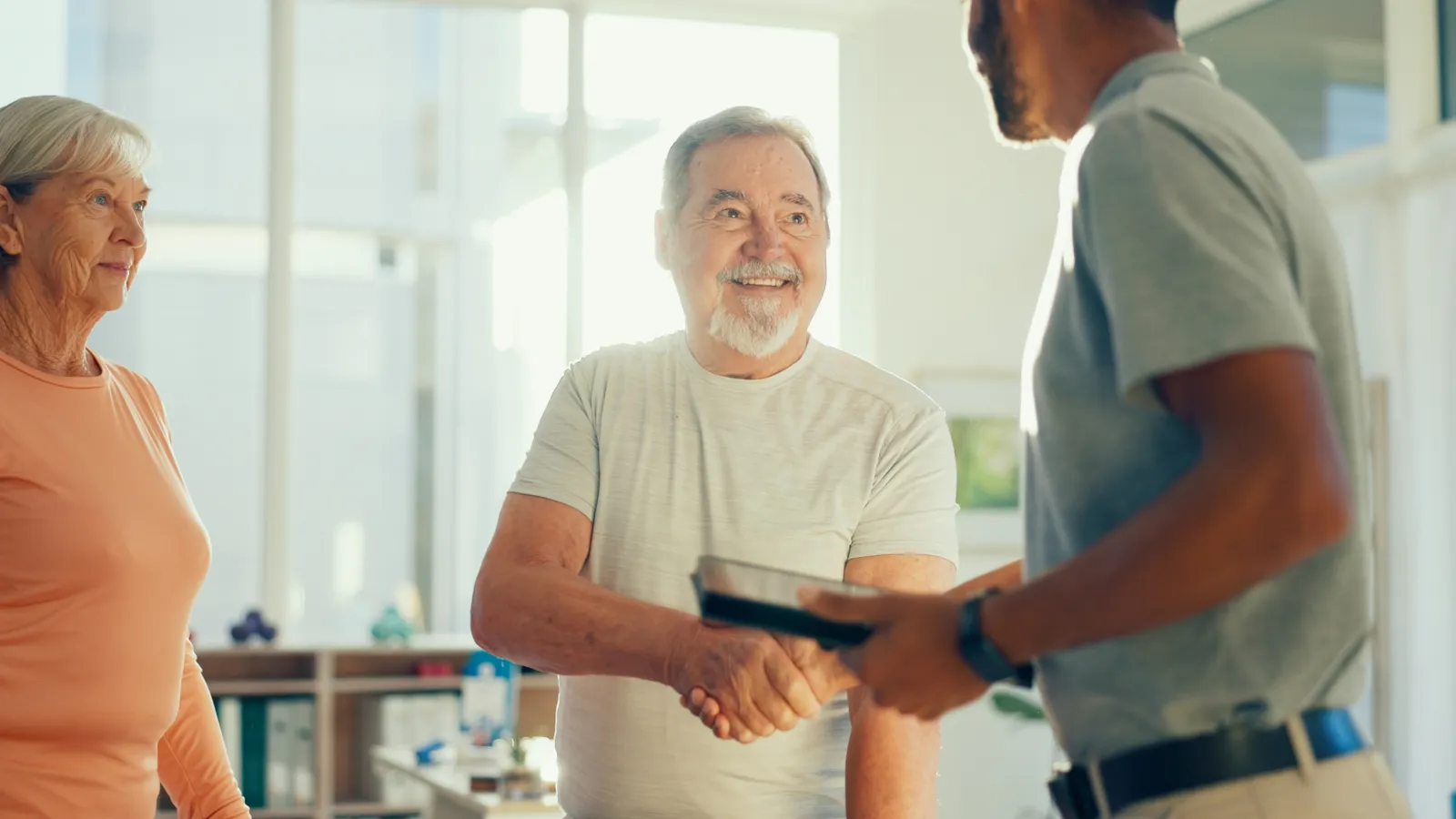 Senior man smiling and shaking hands with a younger man in a bright office with a senior woman observing.