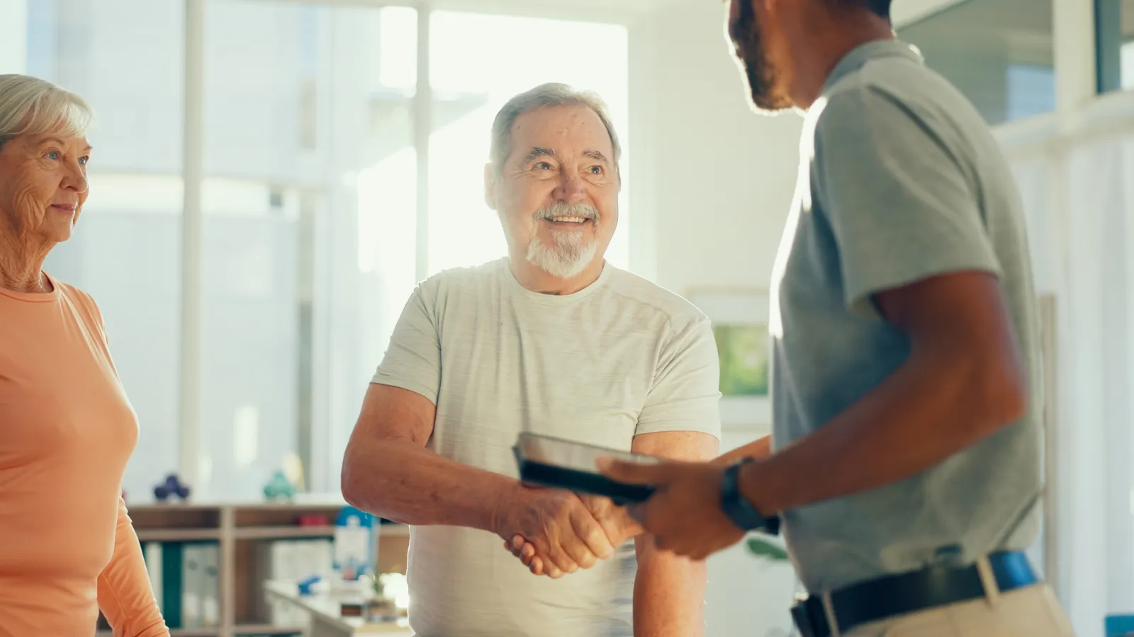 Senior man smiling and shaking hands with a younger man in a bright office with a senior woman observing.
