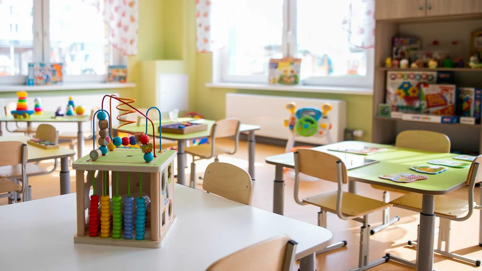 Bright preschool classroom with wooden toys, tables, chairs, and colorful educational materials near windows.