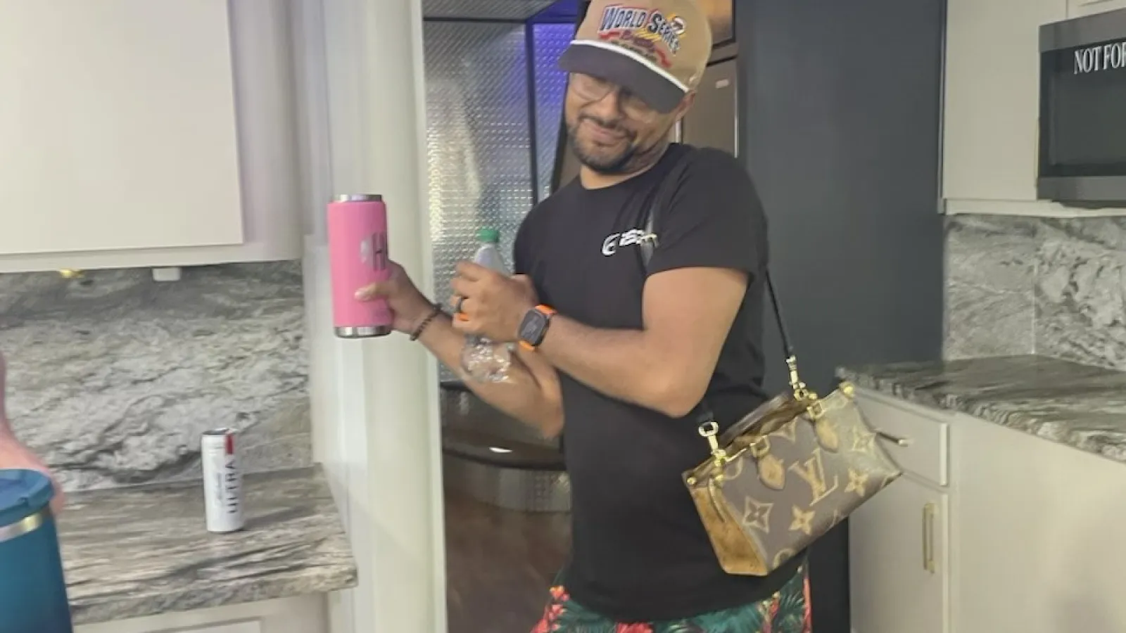 Man in tropical shorts and black t-shirt posing with a pink tumbler and Louis Vuitton purse in a modern kitchen.