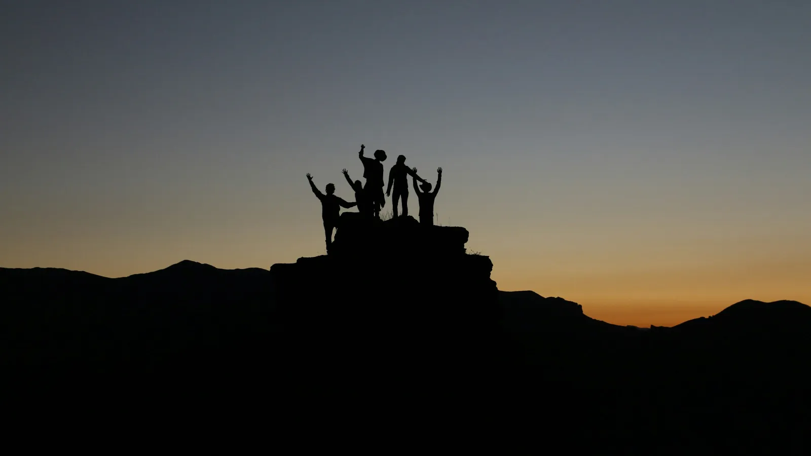 Silhouettes of five people cheering on a rock formation at sunset with mountains in the background.