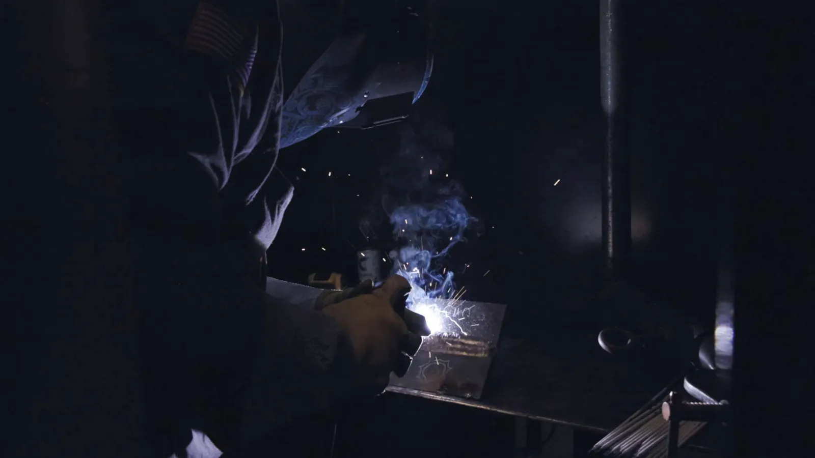 Welder wearing protective gear sparks and smoke illuminate dark workshop during metal welding process.