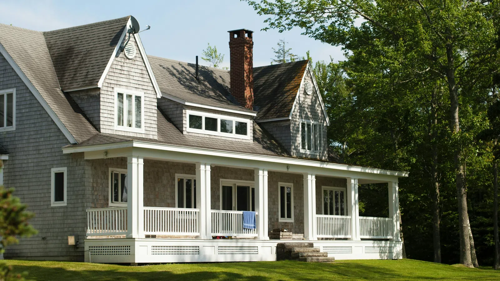 Large gray house with a wraparound porch and chimney surrounded by green lawn and trees on a sunny day.