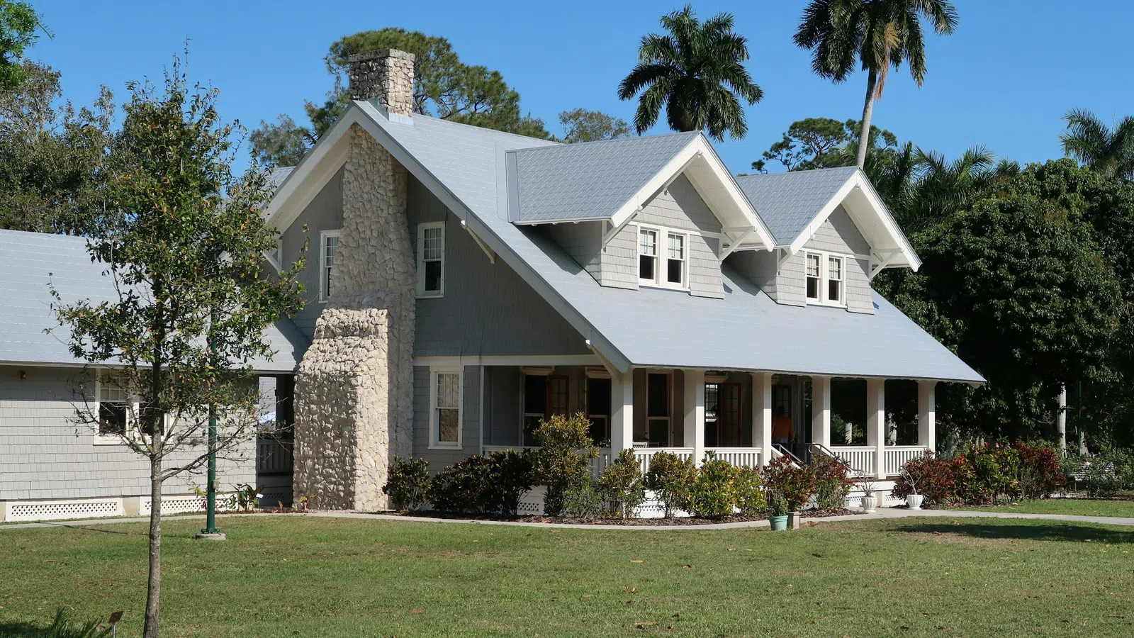 Large gray house with stone chimney, front porch, and lush greenery under a clear blue sky