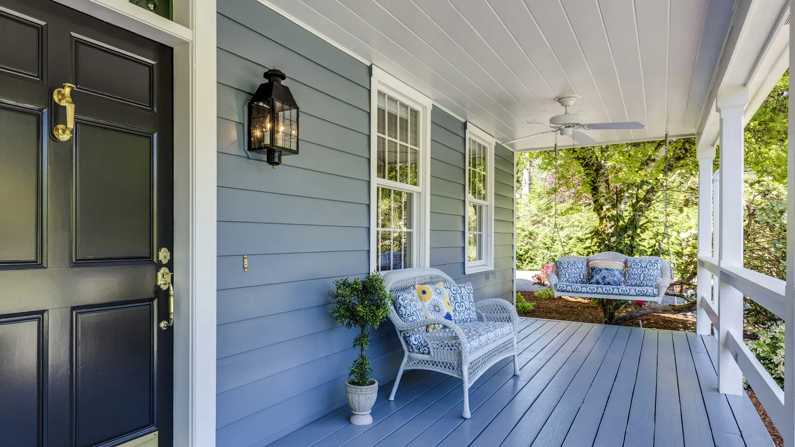 Cozy blue front porch with black door, white wicker chair, ceiling fan, and swing overlooking garden.
