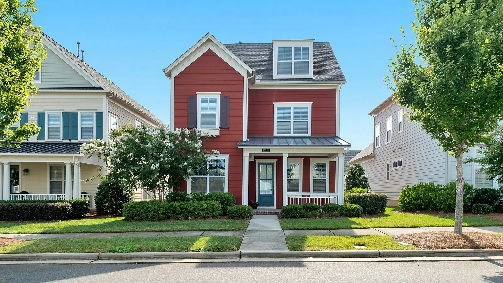 Two-story red house with white trim and black shutters on a sunny day with green trees and lawn.