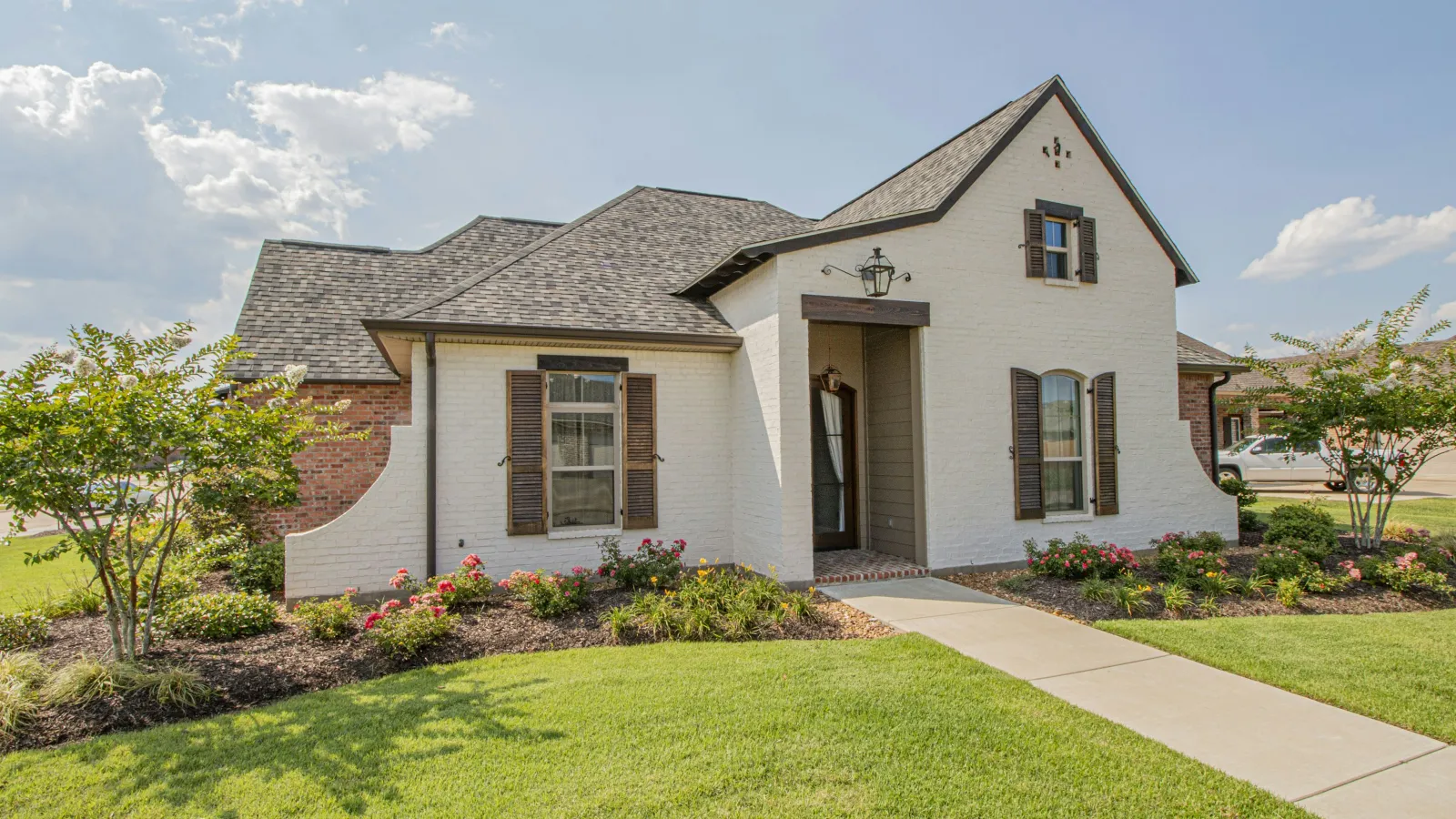 Single-story white brick house with gray roof, brown shutters, manicured lawn, and flower beds under a blue sky.