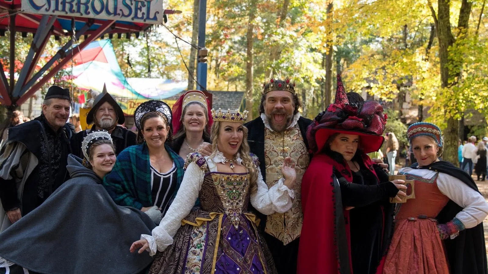 Group of people in elaborate Renaissance costumes posing happily in front of a carousel at a fair under autumn trees.