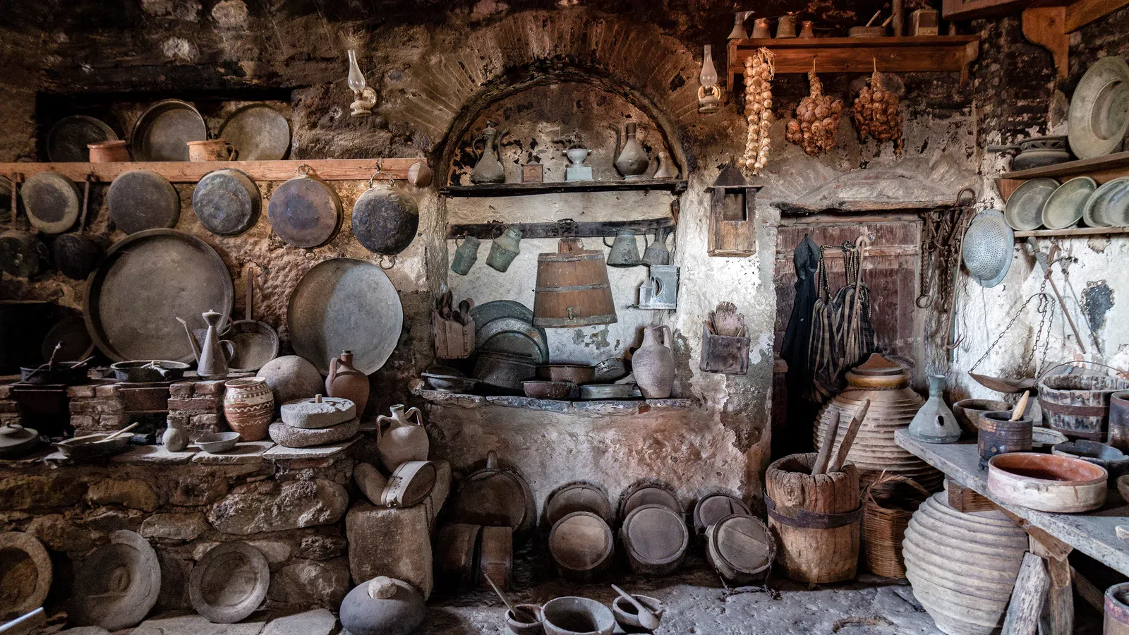 Rustic old kitchen with stone walls, shelves of clay pots, pans, jugs, and vintage kitchenware.