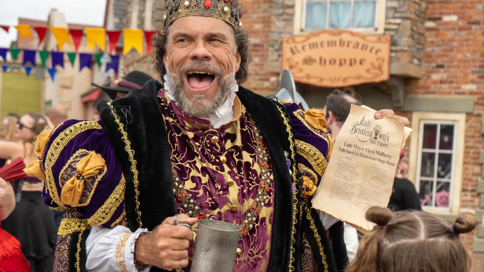Man dressed as a medieval king with crown and robe holding a mug and a parchment at a Renaissance fair.