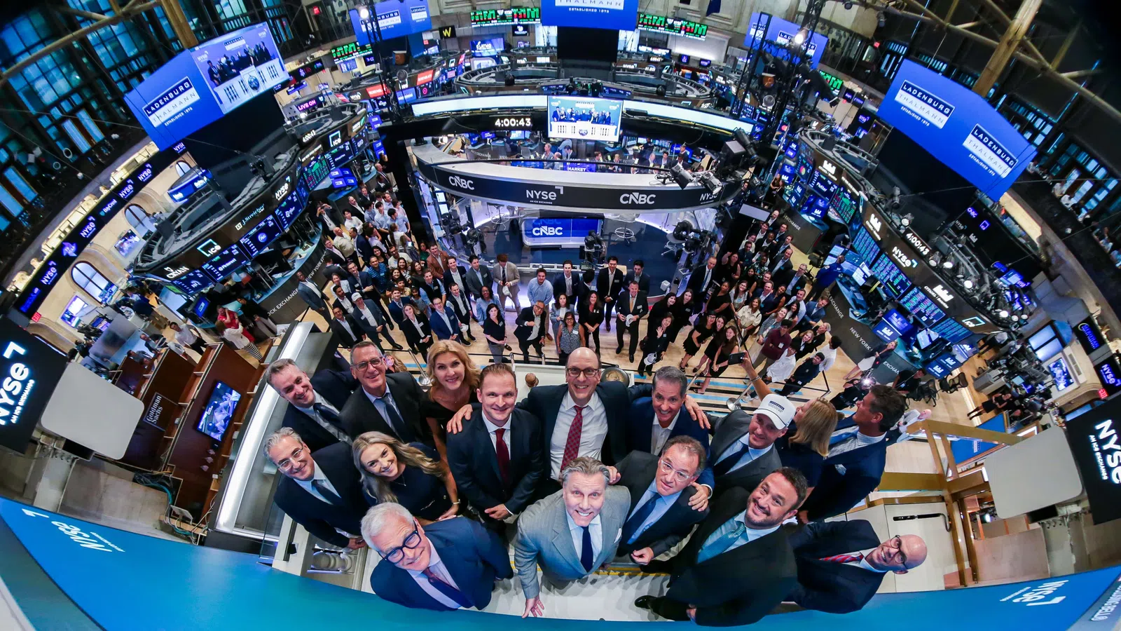 Group of business professionals gathered on the New York Stock Exchange trading floor under large screens.
