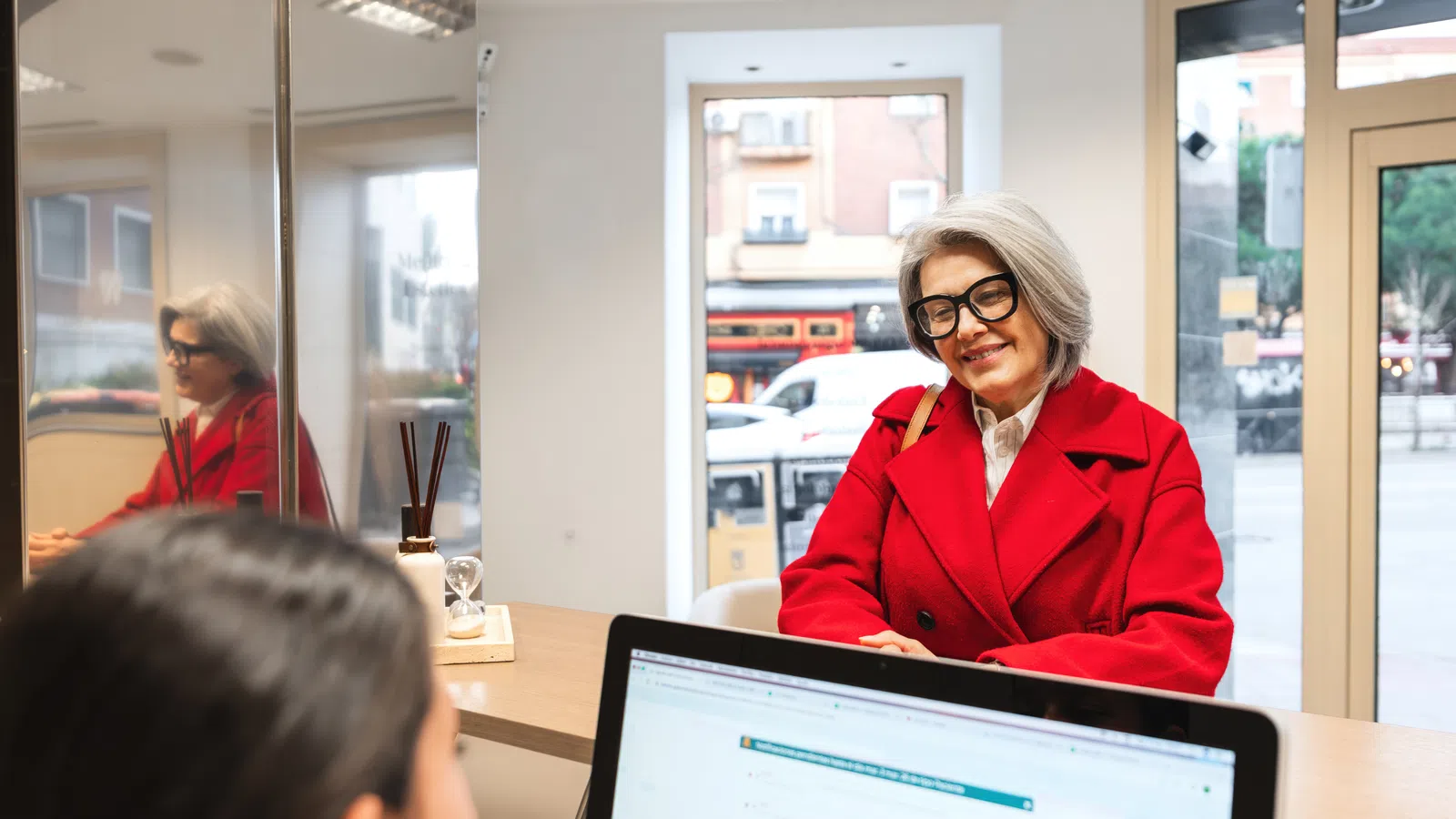 Senior woman in red coat and glasses smiling during consultation at office reception desk.
